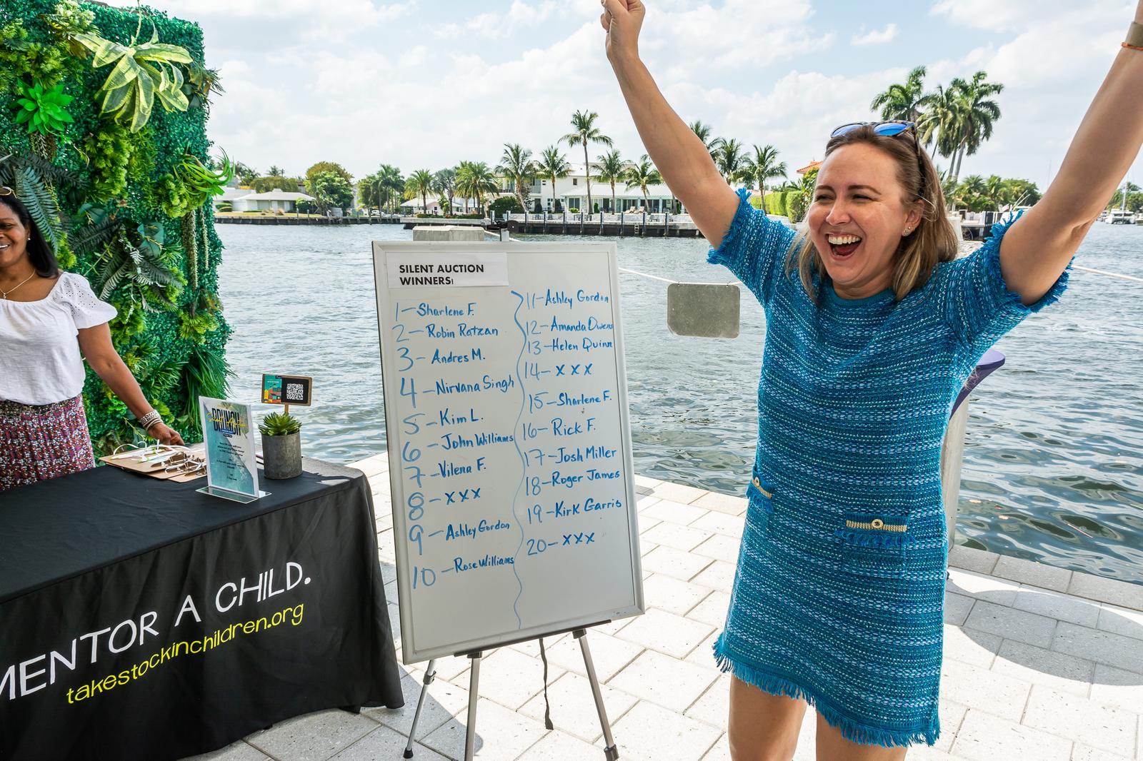 A volunteer celebrating beside a Mentor a Child sign-up board at a waterfront event