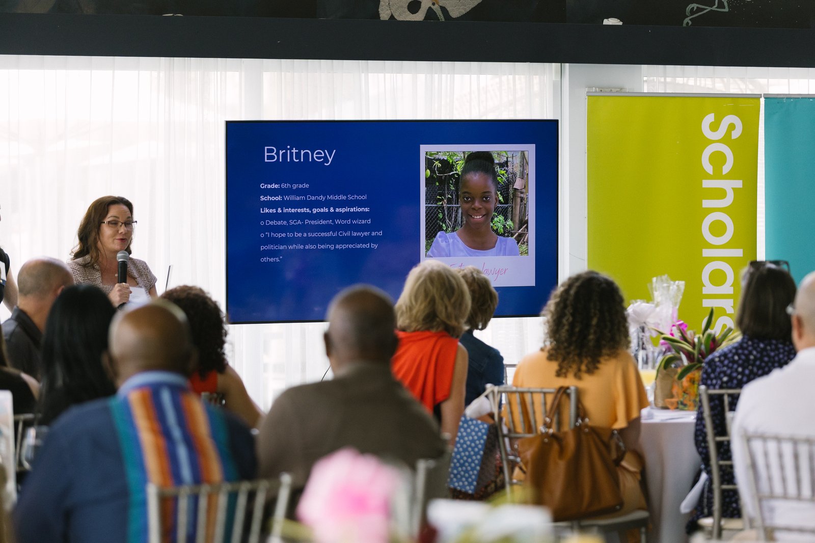 A room of guests watching a scholar's story presented on screen beside the Scholarships banner